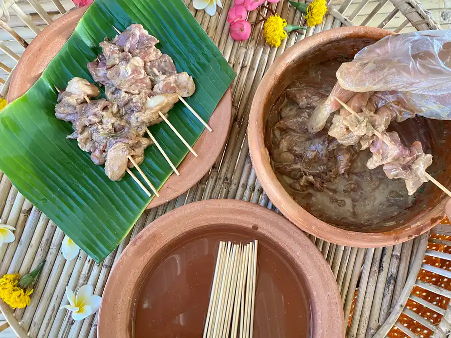 Skewer in the pork on bamboo skewers. There are 3 clay bowl: one with bamboo skewers and water, one with marinated pork, and one with raw pork on skewers.
