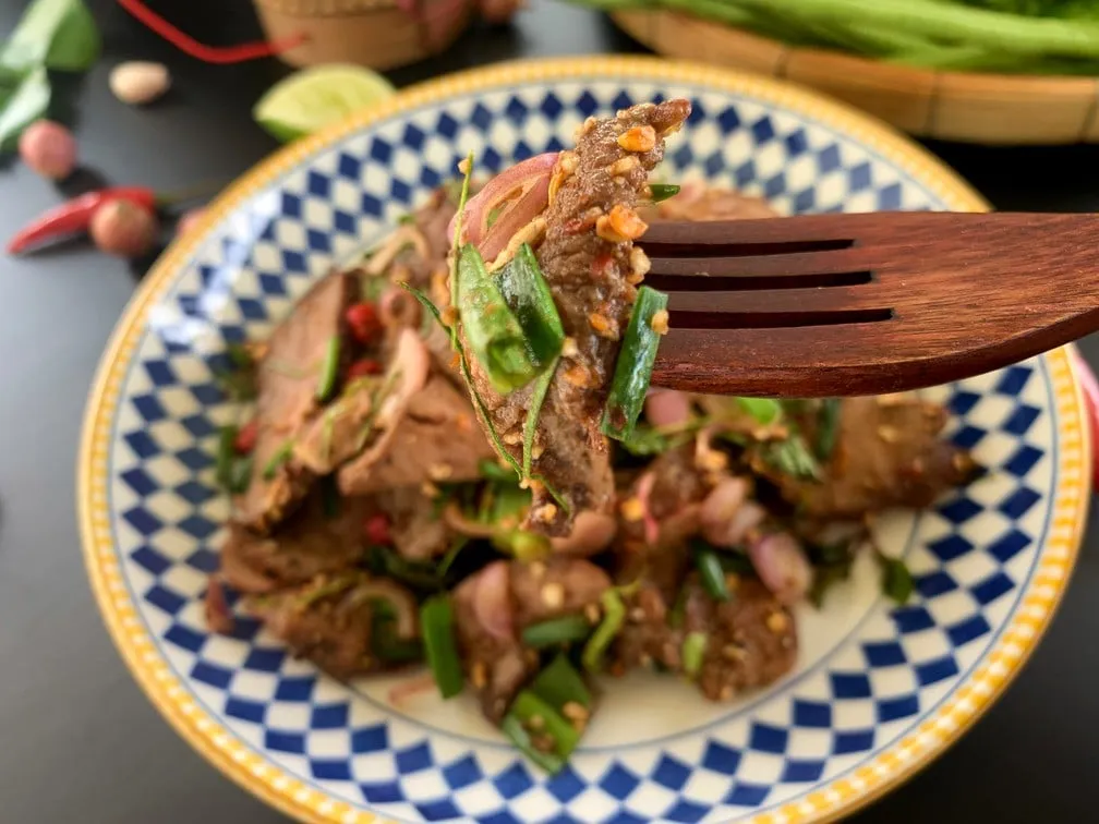 Close-up of Thai beef salad with a wooden fork, lifting a slice of beef.