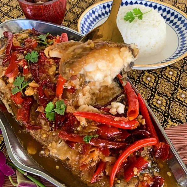 Holding a piece of Thai crispy fish with tamarind sauce in a golden spoon above a plate filled with fish, with white rice and a small mortar and pestle in the background.