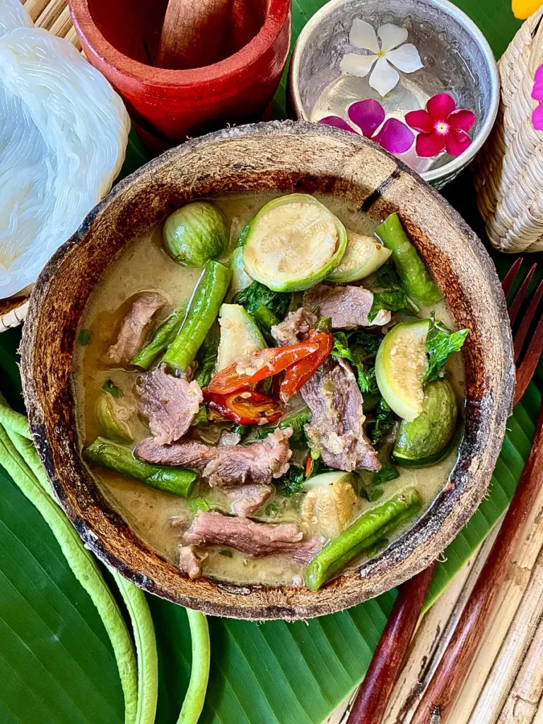 Authentic Thai green curry pork, known as gaeng keow wan, served in a coconut shell bowl with vegetables on a banana leaf.