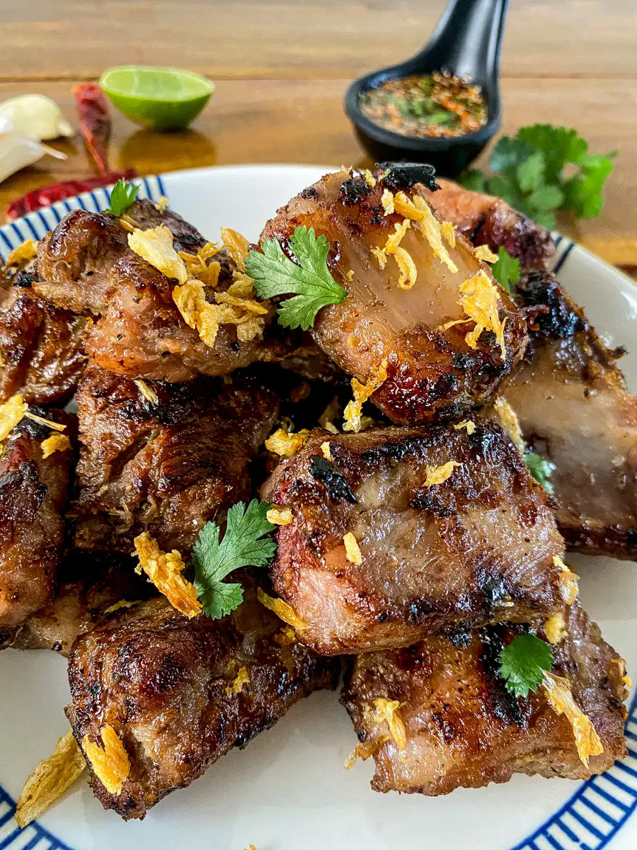 Plate of grilled Thai pork ribs garnished with crispy fried garlic and cilantro, served with a spicy dipping sauce in the background.