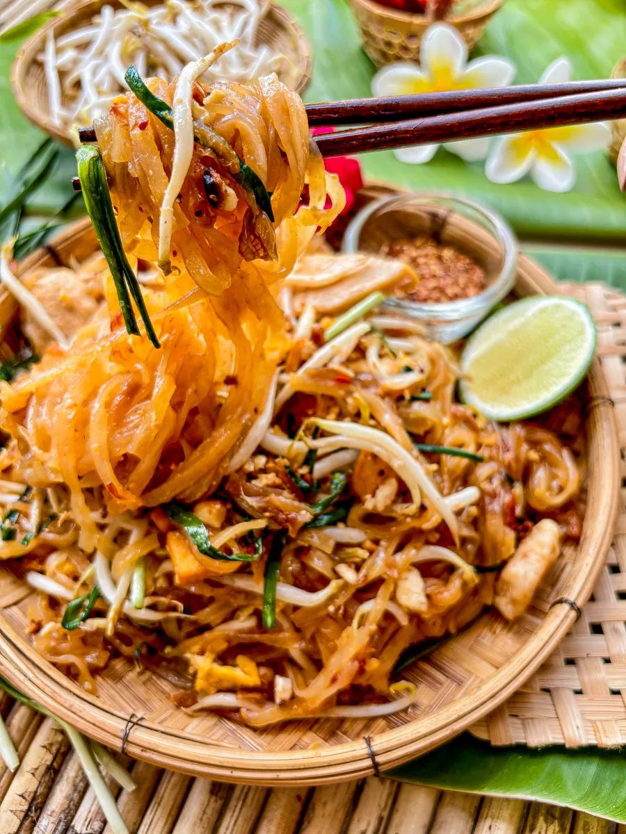 Chicken pad Thai served in a woven bamboo plate with lime, chili flakes, and fresh bean sprouts on the side.