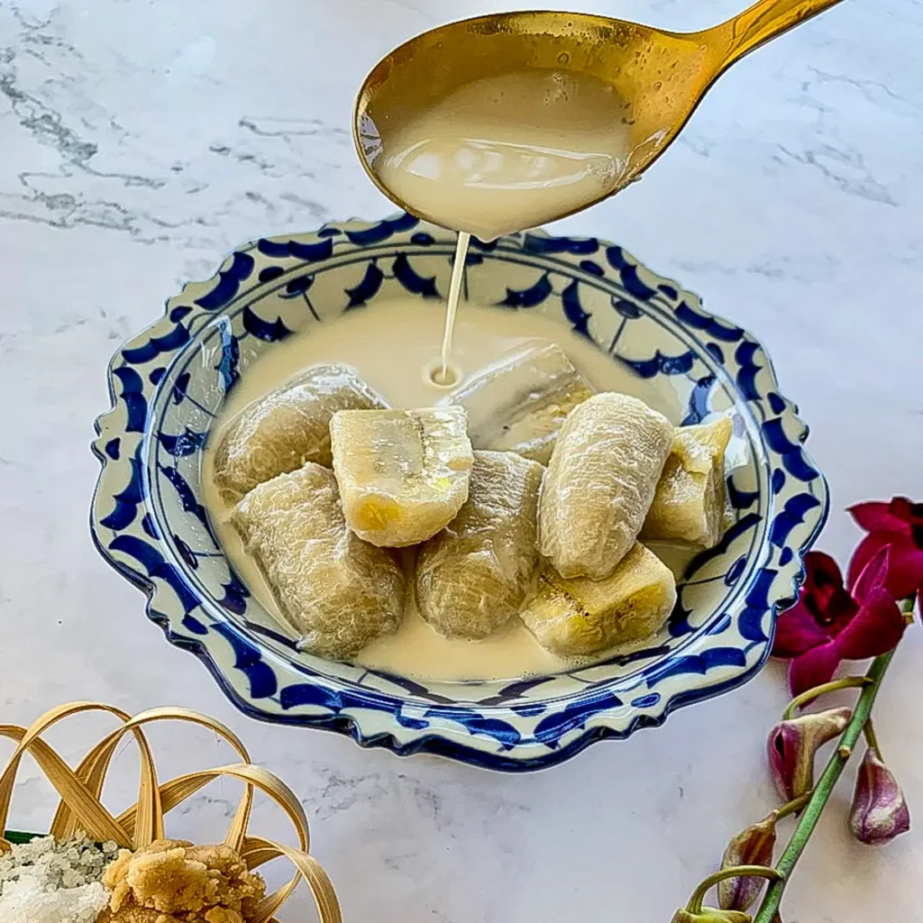 Banana in coconut milk with creamy syrup being poured over sliced bananas in a decorative bowl, garnished with orchids on the side.