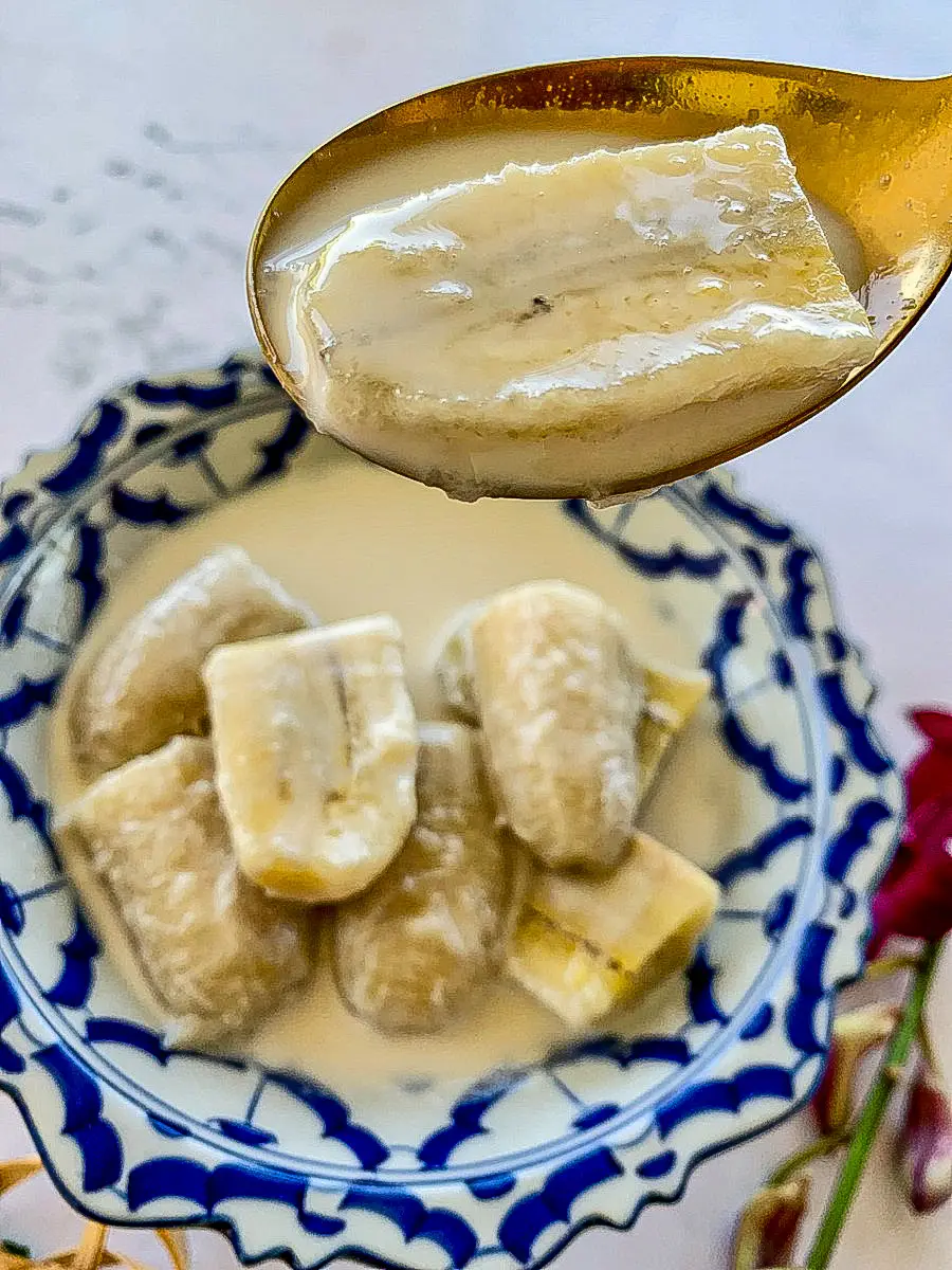 Traditional Thai dessert of bananas simmered in sweet coconut milk, served in a patterned blue and white bowl with a spoonful lifted.