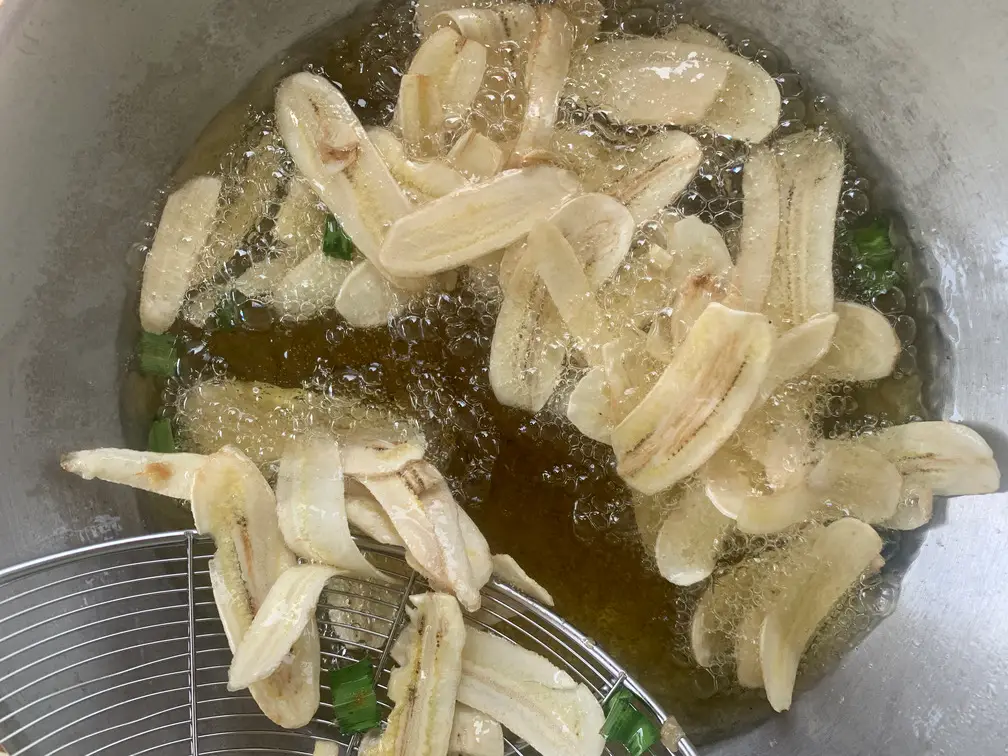 Long strips of banana chips deep-frying in a pot of golden oil alongside green pieces of aromatic pandan leaves.