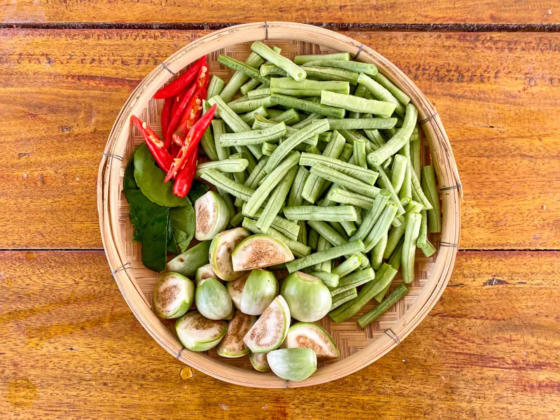 A bamboo tray with chopped yardlong beans, sliced Thai eggplants, red chilies, and kaffir lime leaves.