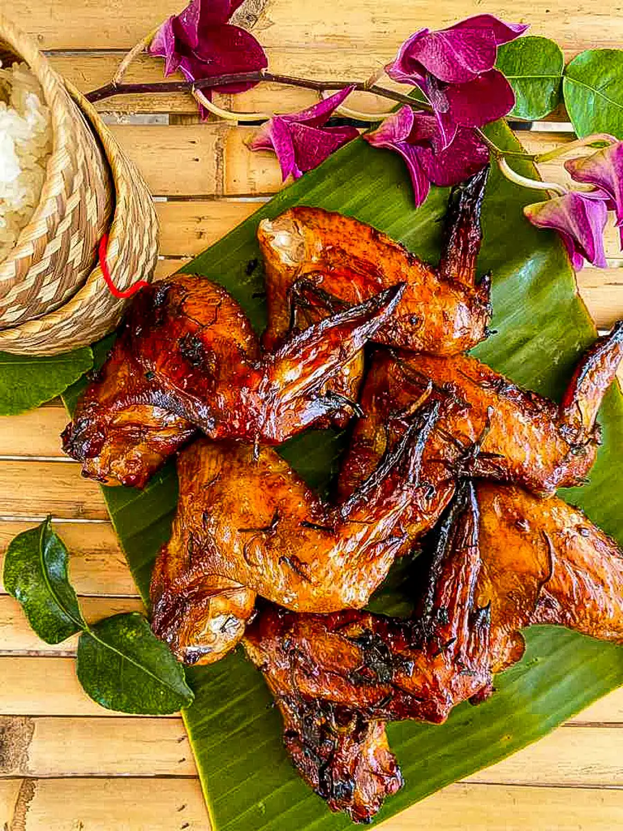Gai yang, grilled Thai chicken wings on a banana leaf with sticky rice and purple orchids on a bamboo table.