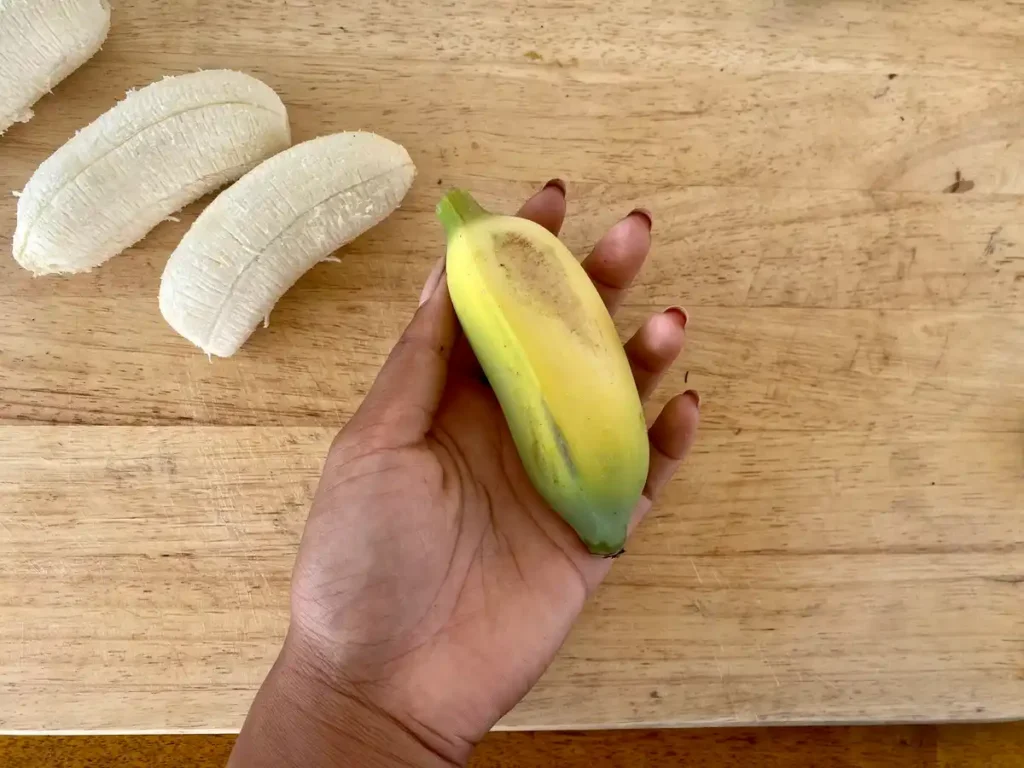 Hand holding an unpeeled Thai banana (kluai namwa) over a wooden cutting board with 2 more peeled bananas next to it.