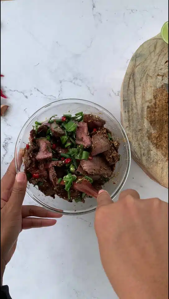Mixing grilled nam tok beef slices with fresh herbs, spices, and sauces in a glass bowl.