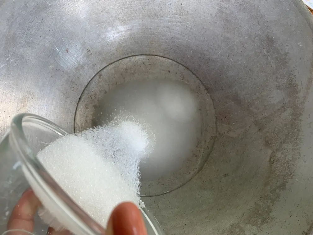 Pouring white granulated sugar from a glass bowl into a metal pan with water to start the sugar-coating syrup.