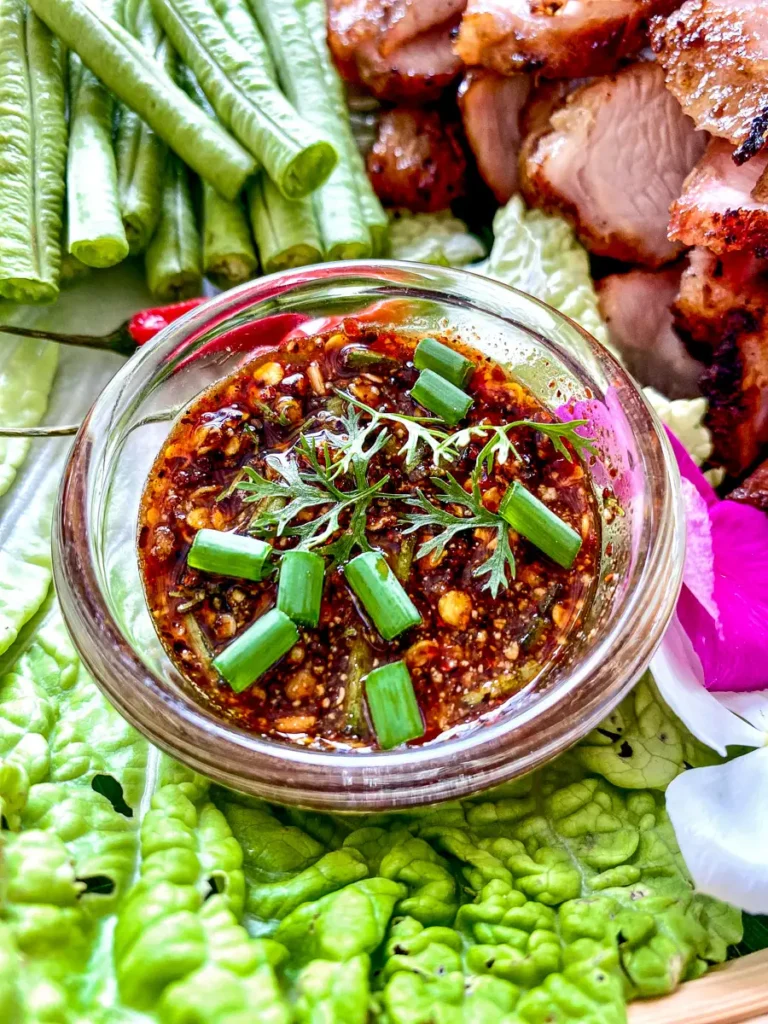 Nam jim jaew sauce in a small bowl topped with herbs and green onions, served with grilled pork, long beans, and cabbage on the side.