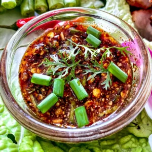 Thai nam jim jaew dipping sauce with chili flakes, toasted rice powder, green onions, and herbs, served in a glass bowl on fresh lettuce.