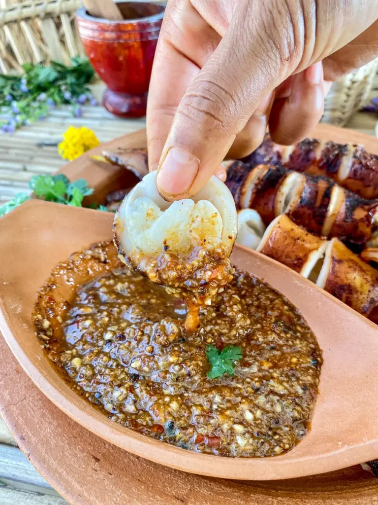 Thai-style grilled squid slice dipped into a bold, chunky nam jim seafood sauce, with garlic, chili, and cilantro in a clay bowl.