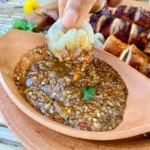 Close-up of grilled squid being dipped into chunky Thai nam jim seafood sauce made with garlic, lime, and chili, served in a terracotta dish.
