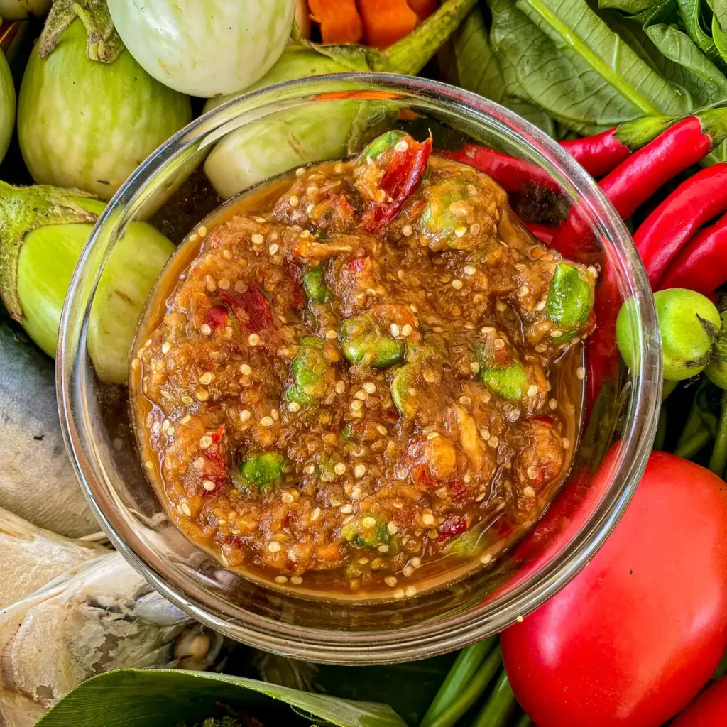 Close-up of nam prik kapi in a sauce cup. Fresh vegetables, chilies, egg, and mackerel are surrounding the chili dip.