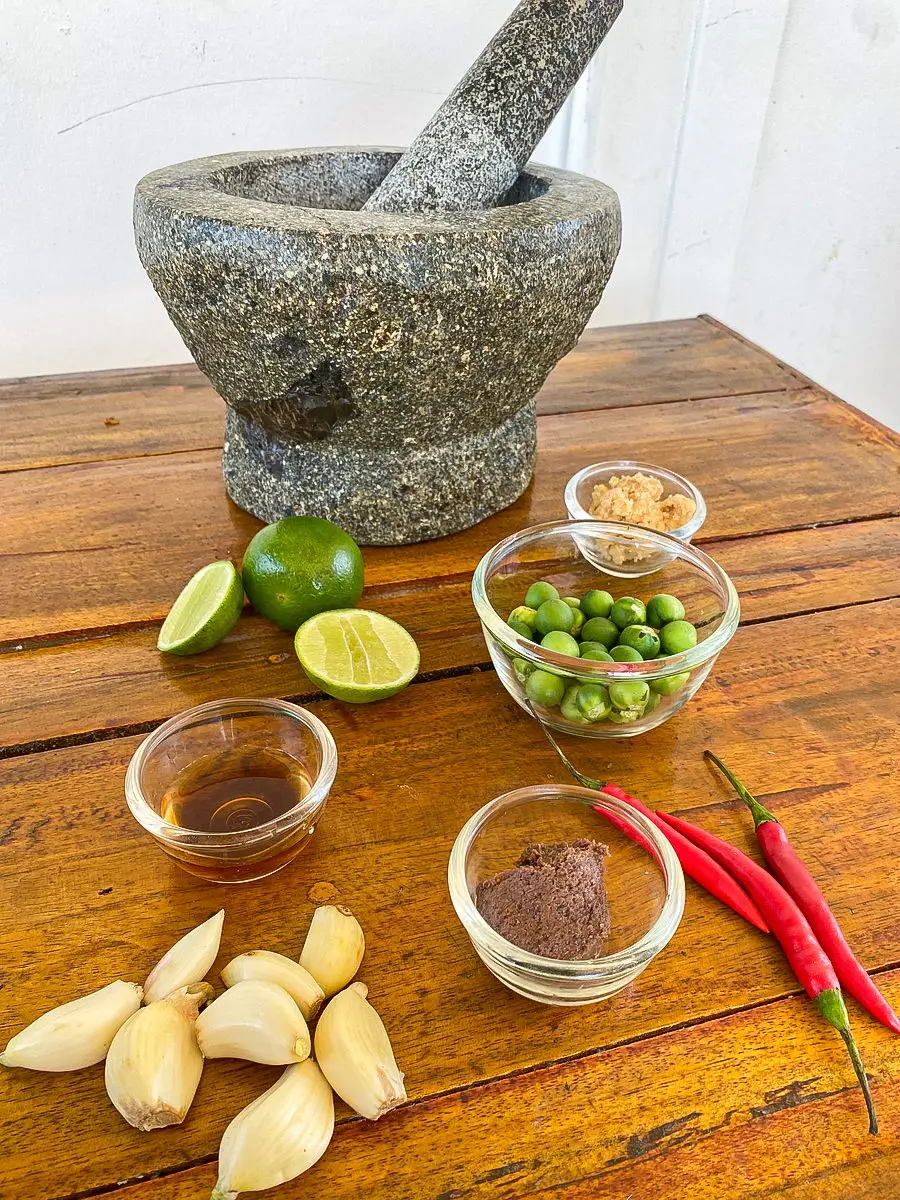 Nam prik kapi ingredients displayed on a wooden table with a granite mortar and pestle; shrimp paste, pea eggplants, garlic, lime, fish sauce, palm sugar, and fresh chili peppers.