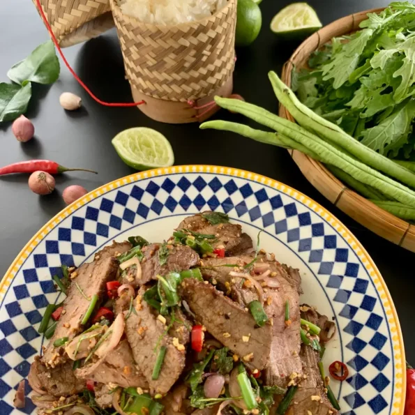A plate of nam tok beef garnished with fresh herbs, alongside sticky rice, red chili peppers, and lime wedges, set on a black surface.