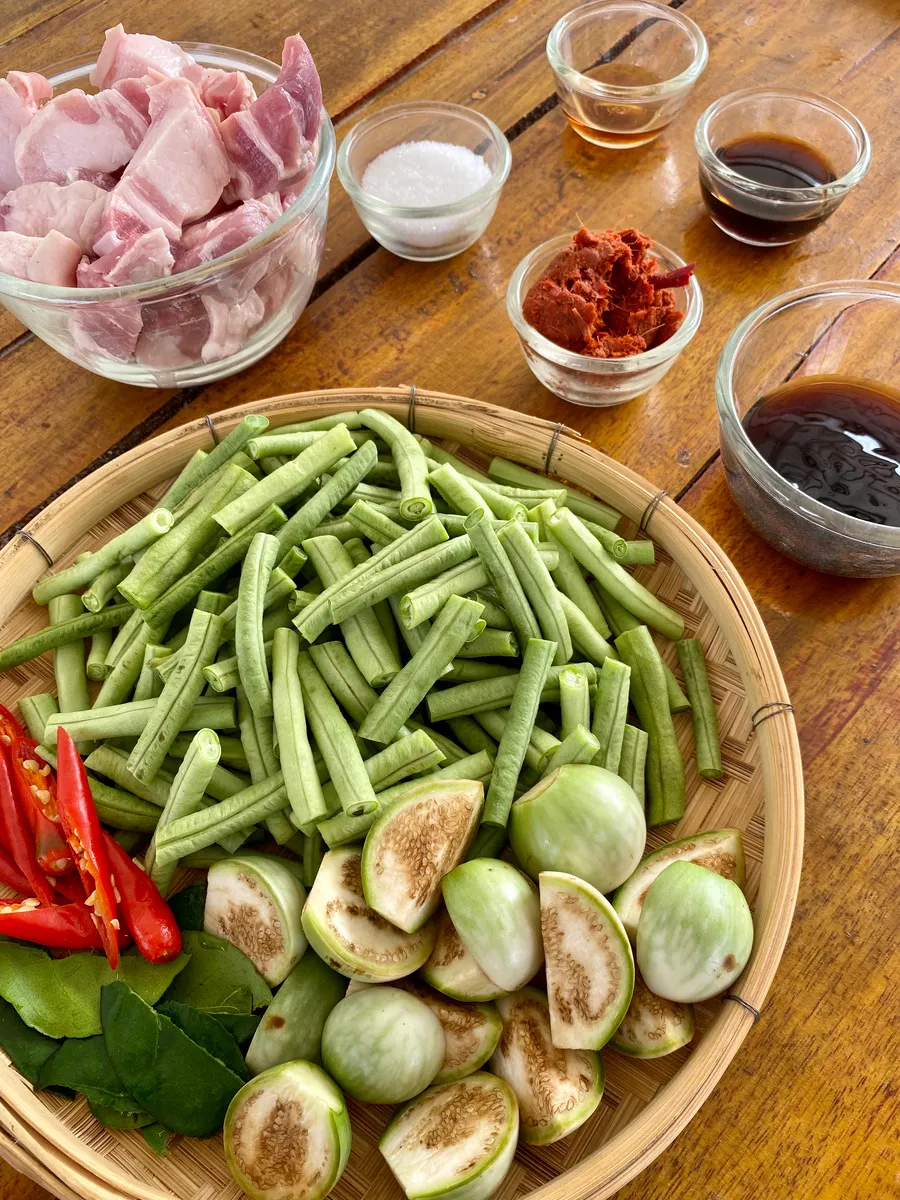 A wooden table topped with a bamboo tray of chopped yardlong beans, halved Thai eggplants, red chilies, and small bowls of pork belly and stir-fry seasonings.