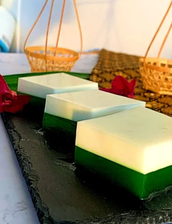 Three square pieces of vibrant green pandan coconut jelly dessert elegantly displayed on a black serving dish, with traditional Thai decorations adorning the background.