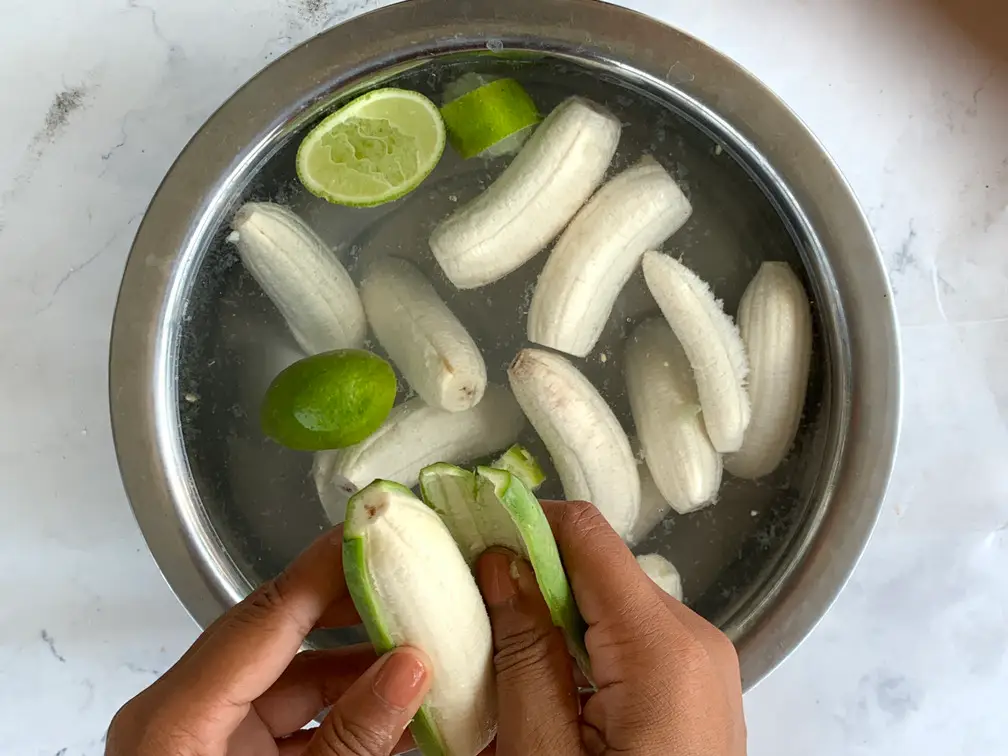 A person peeling green, unripe Thai bananas over a silver bowl of water filled with fresh lime halves and peels to prevent browning.