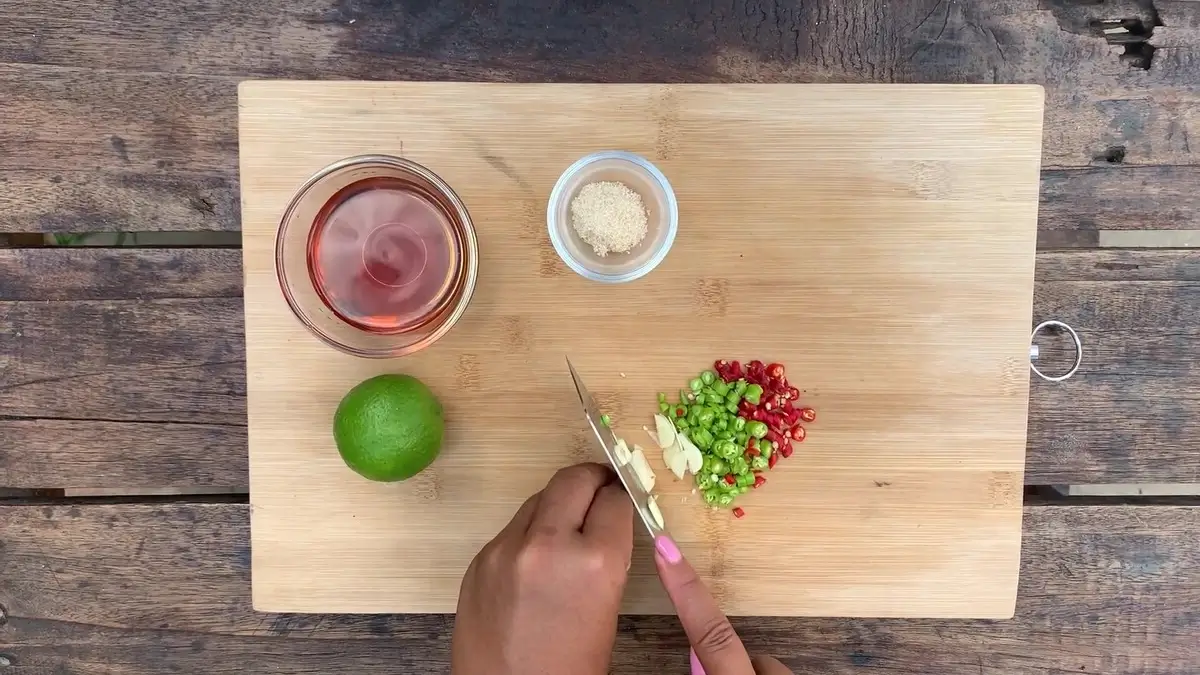 Hands slicing fresh chilies and garlic on a wooden chopping board, with fish sauce, lime, and sugar ready.