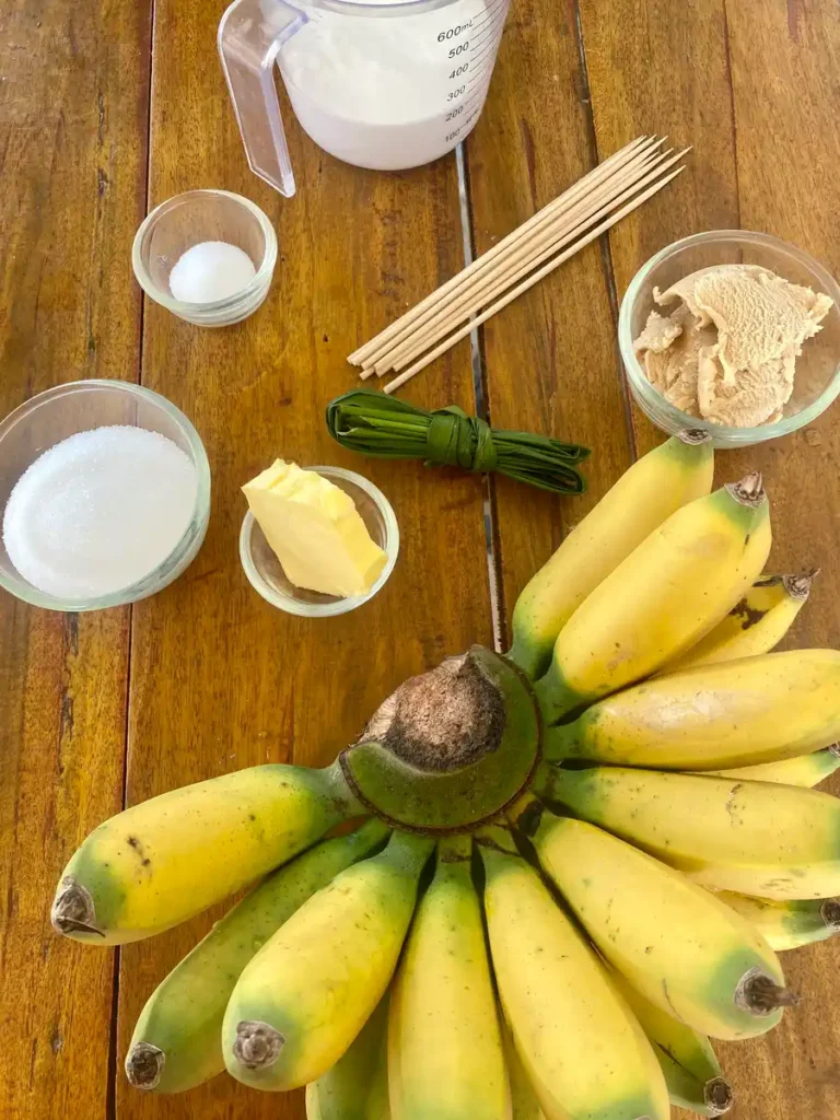 Thai bananas, butter, white sugar, palm sugar, salt, skewers, pandan leaves, coconut milk on a wooden background.