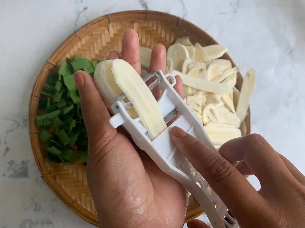 Using a white vegetable peeler to slice a firm, green banana into long, thin uniform strips over a woven bamboo tray.