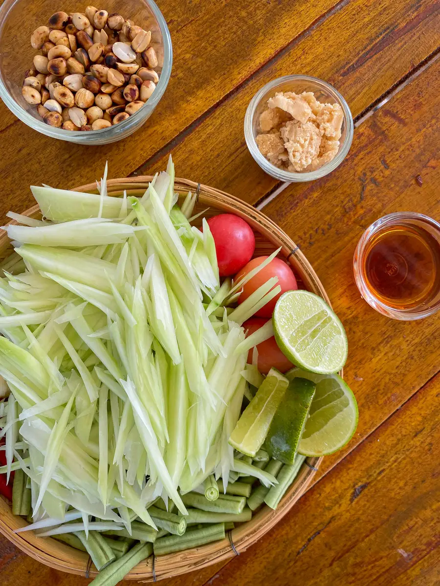Green papaya, tomatoes, lime, palm sugar, peanuts, and fish sauce on a wooden table.