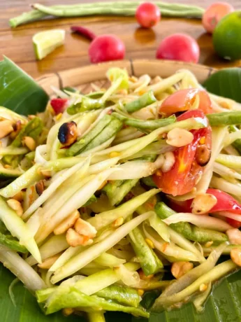 Close-up image of Som Tum Thai, featuring shredded papaya, sliced tomatoes, lime, and peanuts, artfully presented on a banana leaf in a bamboo dish.