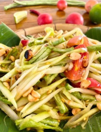 Close-up image of Som Tum Thai, featuring shredded papaya, sliced tomatoes, lime, and peanuts, artfully presented on a banana leaf in a bamboo dish.