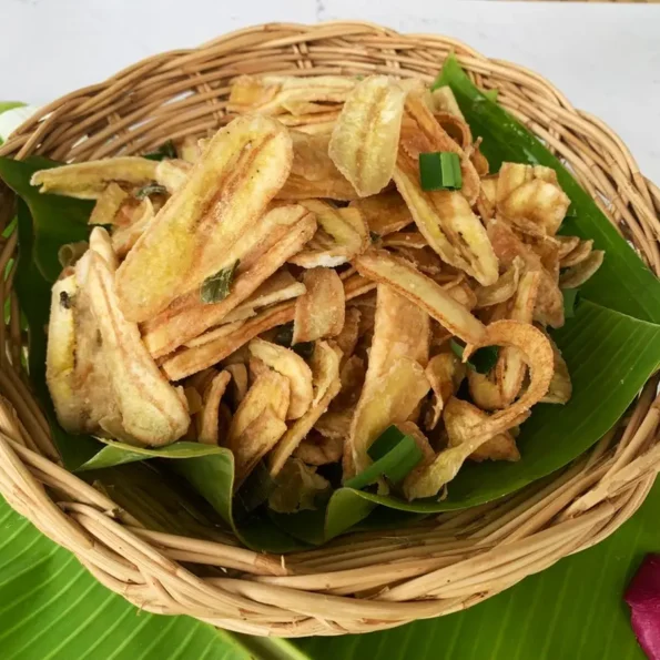 A basket of crispy Thai fried banana chips, served on banana leaves with tropical flowers around.