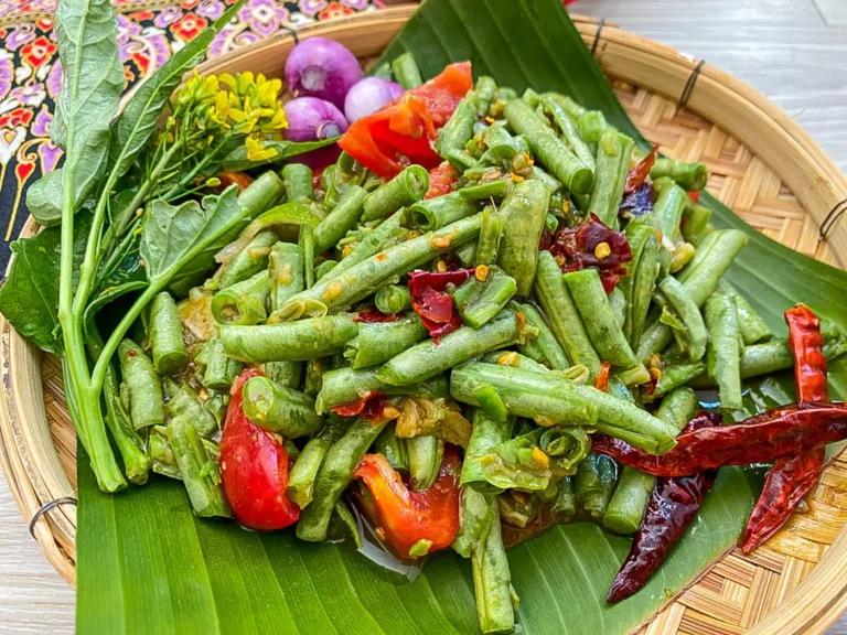 Thai long bean salad with tomatoes and dried chilies, served with raw shallots and yellow flowers on a banana leaf.