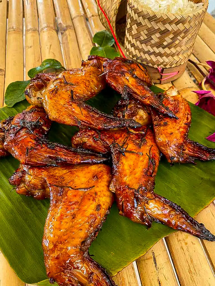 Thai wings with golden, caramelized skin served on banana leaves next to sticky rice in a woven container.