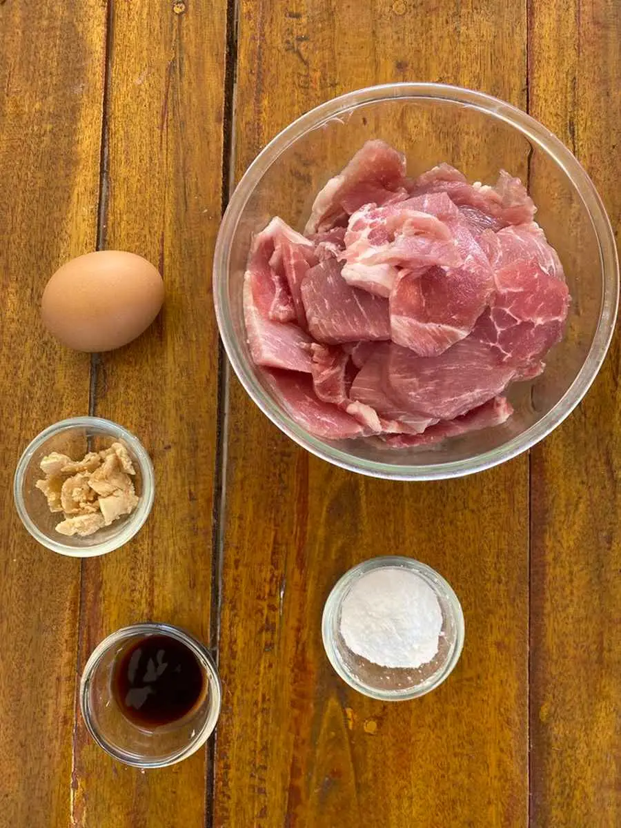 Ingredients for pork marinade: An egg, pork, palm sugar, tapioca starch, and oyster sauce in glass cups.