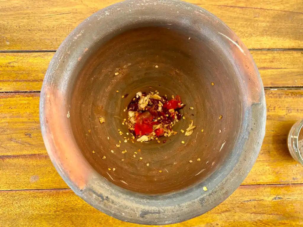 Top-down view of pounded chilies and garlic in a clay mortar on a wooden background.