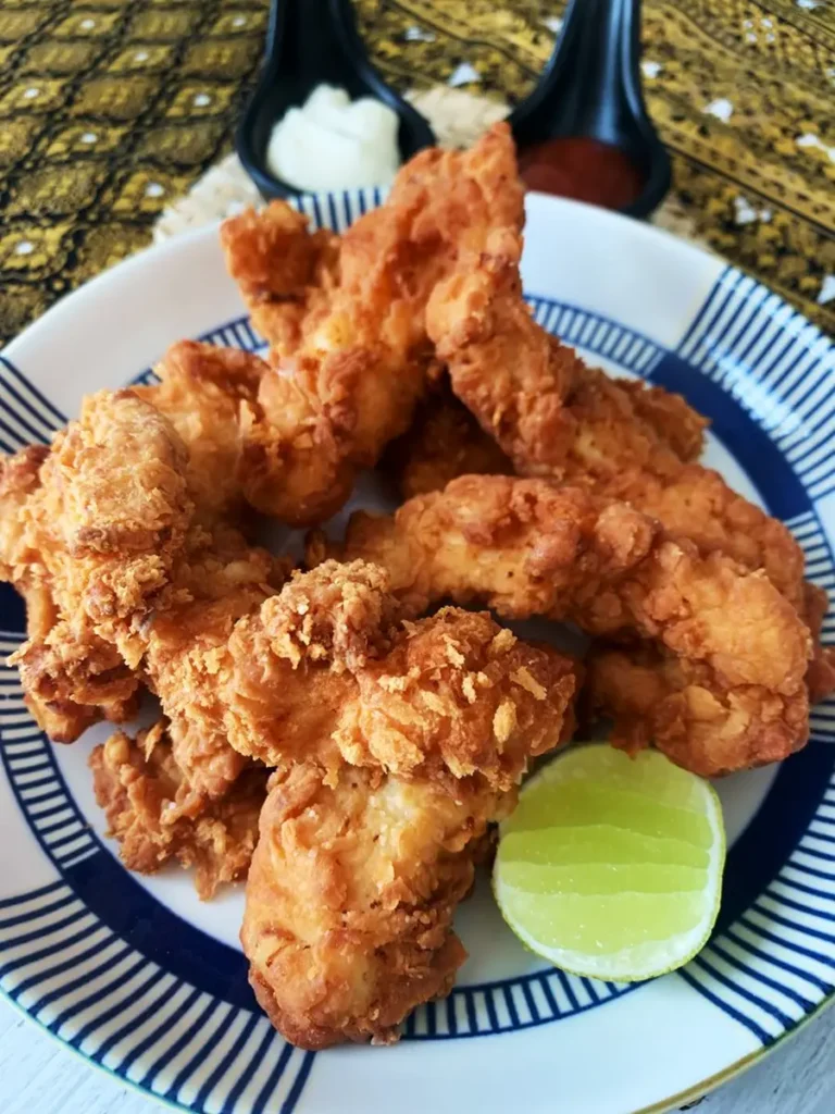 Thai fried chicken tenders on a white dish with a lime wedge. Above it are 2 dipping sauces in black cups; mayonnaise and ketchup.