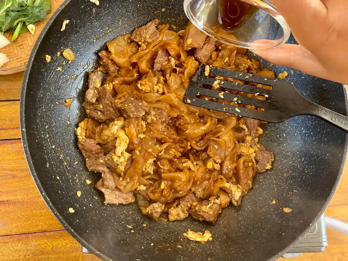 Adding soy sauce mixture to noodles and beef in a wok.
