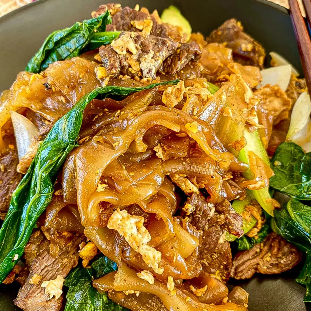Close-up of beef pad see ew showing chewy stir-fried rice noodles, seared beef slices, and wilted Chinese broccoli in a dark bowl.