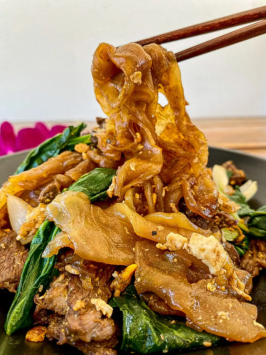 Fresh wide rice noodles stir-fried with beef, Chinese broccoli, and egg, lifted by chopsticks for a close-up view.