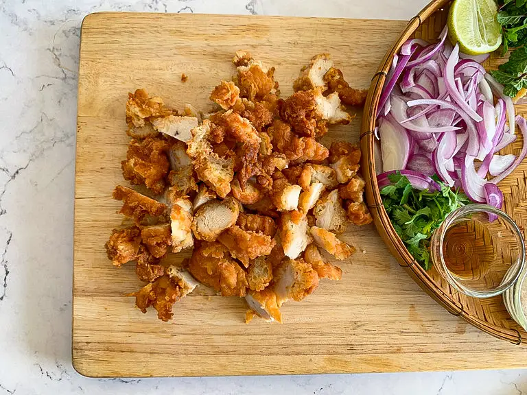 Cut fried chicken pieces on a wooden board with sliced shallots and herbs on the side.