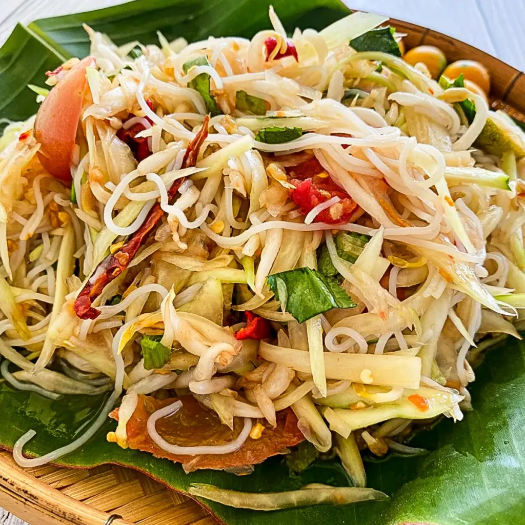 Tam sua, Thai papaya salad with rice vermicelli, fermented fish sauce, tomatoes, chilies, and lime leaves served on banana leaf in a bamboo tray.