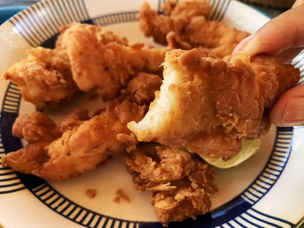 Close-up of fried chicken held in hand, with a bite taken, presented on a white dish alongside more crispy fried chicken.