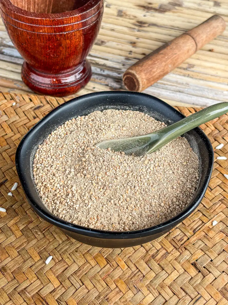 Toasted rice powder (khao khua) in a black bowl with a green spoon, set on a woven mat beside a traditional wooden mortar and pestle.
