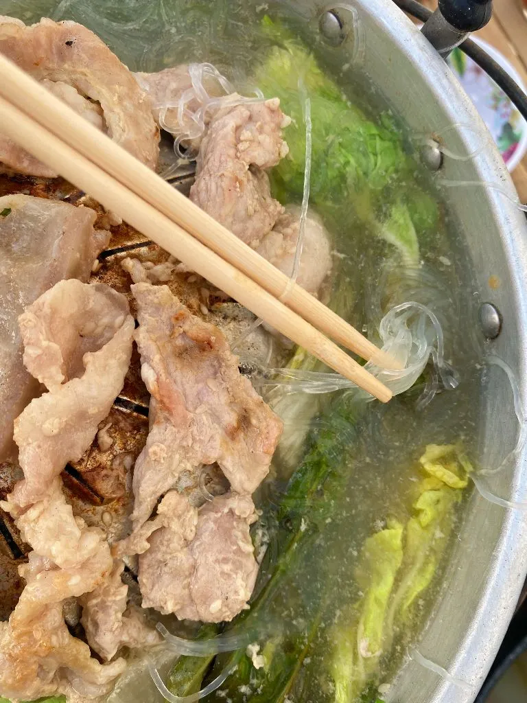 Holding glass noodles with chopsticks in the soup base of mookata, with pork meat on top of the grill.
