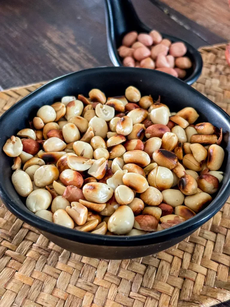 Black bowl filled with dry roasted peanuts, some split open, placed on a woven mat with another small dish of peanuts in the background.