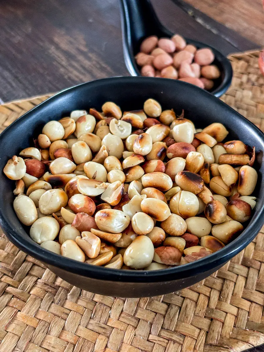 Black bowl filled with dry roasted peanuts, some split open, placed on a woven mat with another small dish of peanuts in the background.