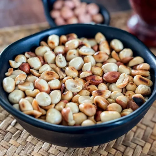 Close-up of dry roasted peanuts in a black bowl on a woven mat, with raw peanuts visible in a spoon-shaped dish behind