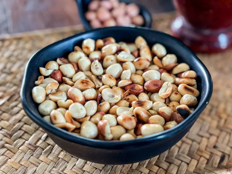 Close-up of dry roasted peanuts in a black bowl on a woven mat, with raw peanuts visible in a spoon-shaped dish behind