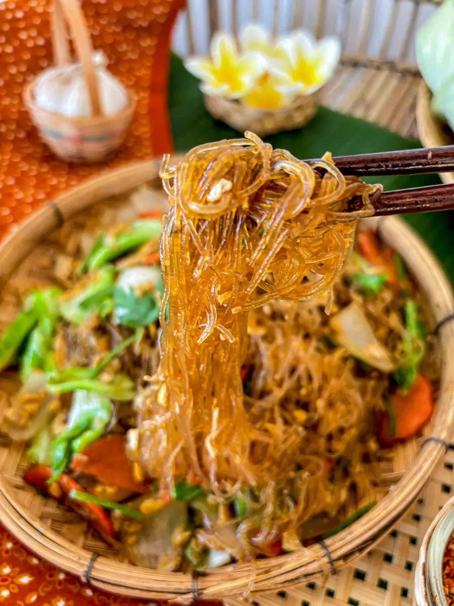 Chopsticks lifting stir-fried glass noodles over a plate of pad woon sen with vegetables.