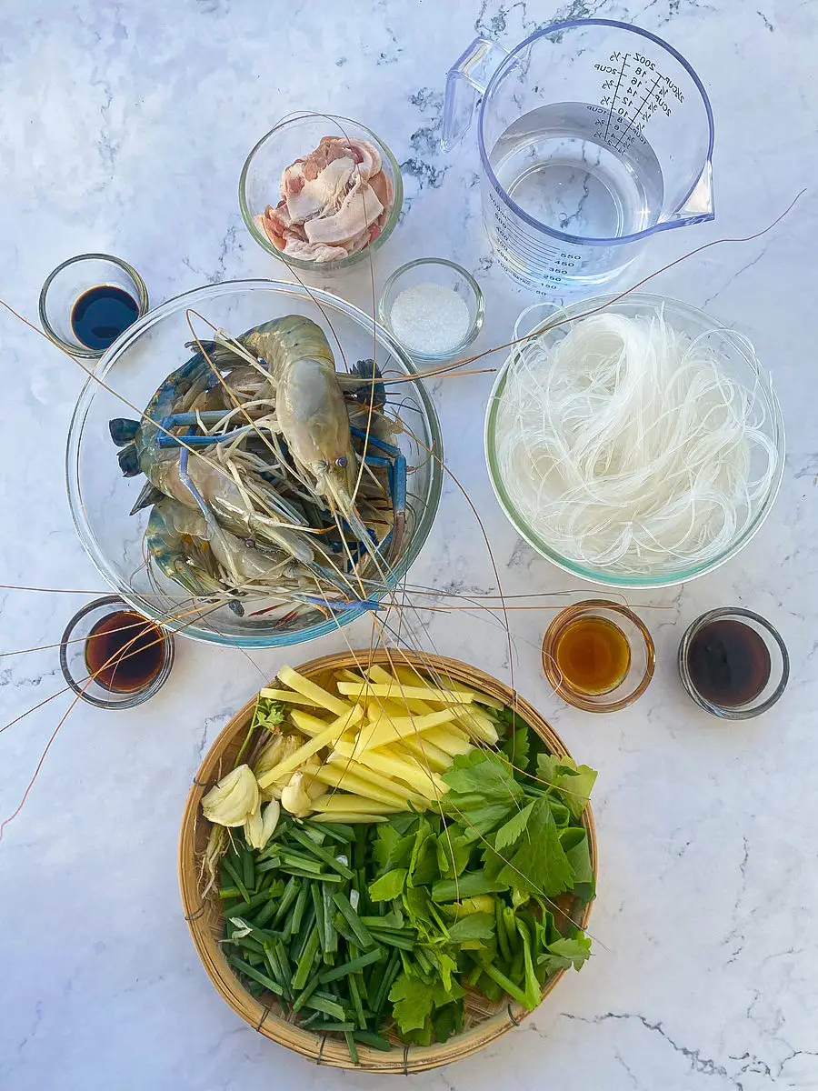 Ingredients for goong ob woonsen including prawns, glass noodles, bacon, ginger, Chinese celery, garlic, and Thai sauces arranged on a table.